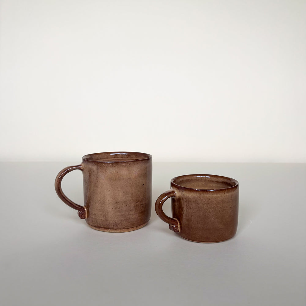 Two brown ceramic mugs on a light cream background with a interisting twirl handle. Pottery Mugs  Handmade in Ireland. Wheel Thrown Pottery and Ceramic. 