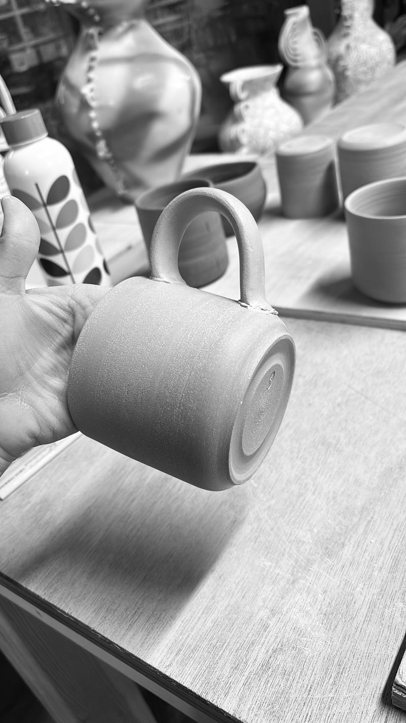 Person holding a ceramic mug attaching the handle with more mugs and bowls in the background on a wooden surface. Handmade pottery Ireland. Handmade Ceramic mug.