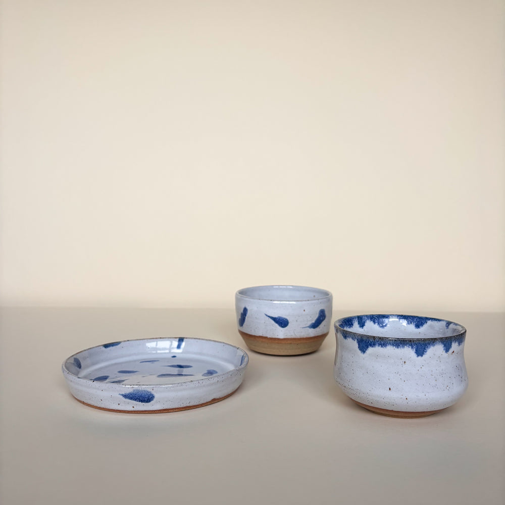 Two white ceramic bowls with blue abstract speckles and a white ceramic plate with blue speckles. On a cream background. Pottery Handmade in Ireland. Wheel Thrown Pottery and Ceramic. Front View. 